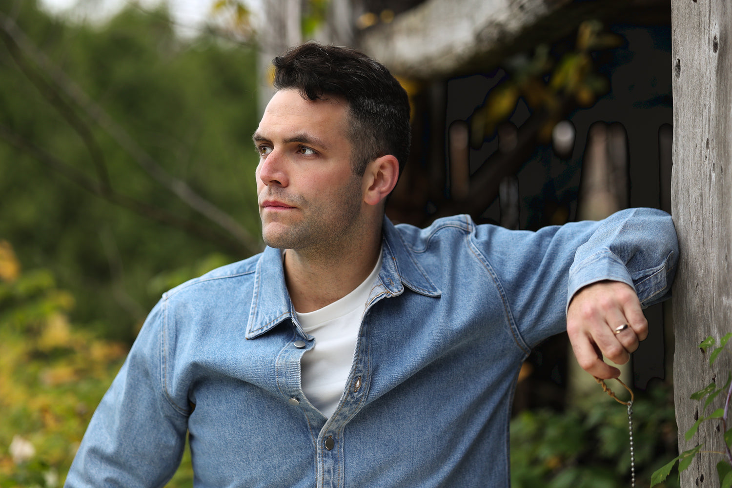 Jeremie Legault, clad in a Canadian tuxedo, strikes a contemplative pose, gazing off to the left with a pensive expression. The rustic charm of the dilapidated barn provides a poetic backdrop, as he channels a thoughtful and introspective mood in this evocative moment.