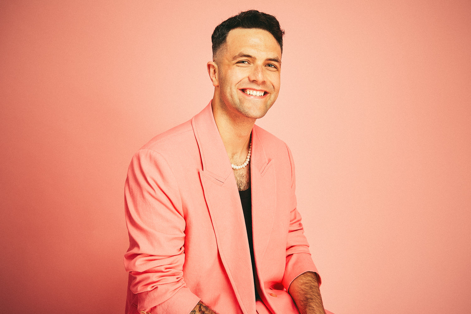 Portrait of Jeremie Legault, a stylish rockstar, smiling broadly while wearing a fashionable pink blazer over a black tank top. He accessorizes with a pearl necklace, exuding confidence and charm. The clean-cut and handsome appearance complements the pink backdrop.