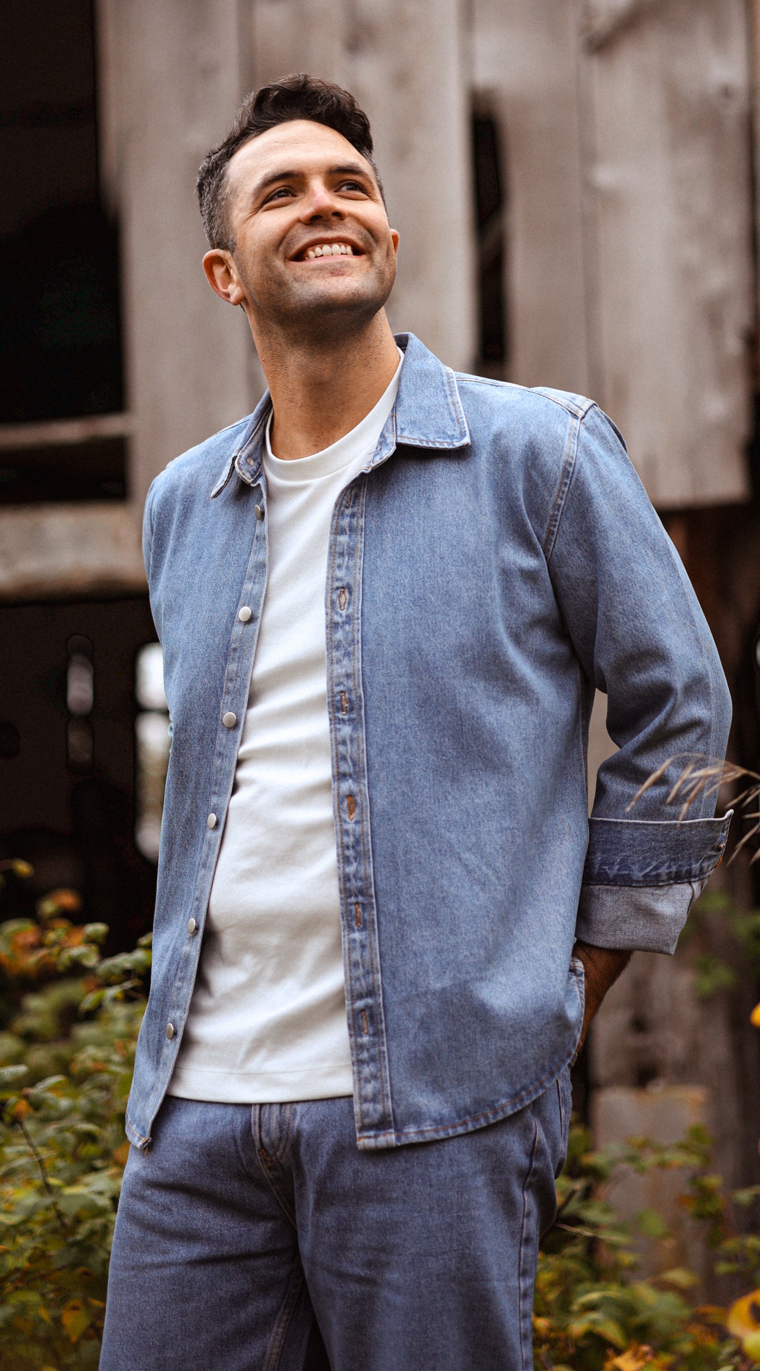 Jeremie Legault, radiating genuine joy, stands confidently in a Canadian tuxedo against the rustic backdrop of a dilapidated barn. His smile reflects the harmonious blend of denim and countryside charm, capturing a moment of authentic Canadian spirit.