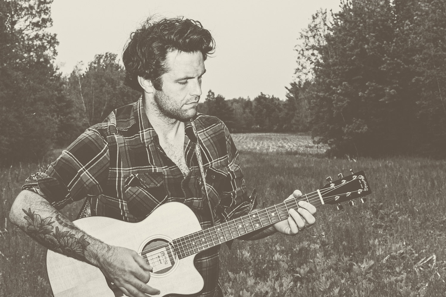 Sepia-toned photo of rockstar Jeremie Legault in a forest clearing, playing a single cutaway Martin acoustic guitar. He is unshaven with medium-length black curly hair and visible tattoos of Canadian flowers on his arms, which are exposed by the rolled-up sleeves of a plaid shirt. His eyes are cast down, concentrating on the D chord he is playing.